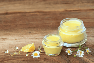 Two glass jars of yellow cannabis-infused body balm with chamomile flowers and shea butter on a wooden surface.