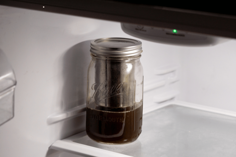 Glass mason jar filled with dark infused oil stored on the bottom shelf of a refrigerator for preservation.