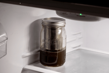 Glass mason jar filled with dark infused oil stored on the bottom shelf of a refrigerator for preservation.