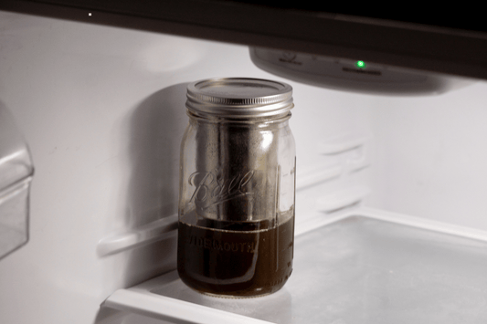 Glass mason jar filled with dark infused oil stored on the bottom shelf of a refrigerator for preservation.