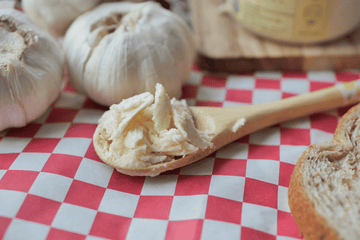 Wooden spoon with garlic butter on checkered red-and-white paper, surrounded by fresh garlic bulbs and toast.